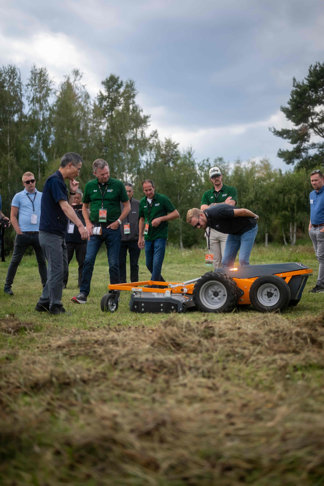 Group inspecting the mowing deck of an orange Raymo carrier in tall grass