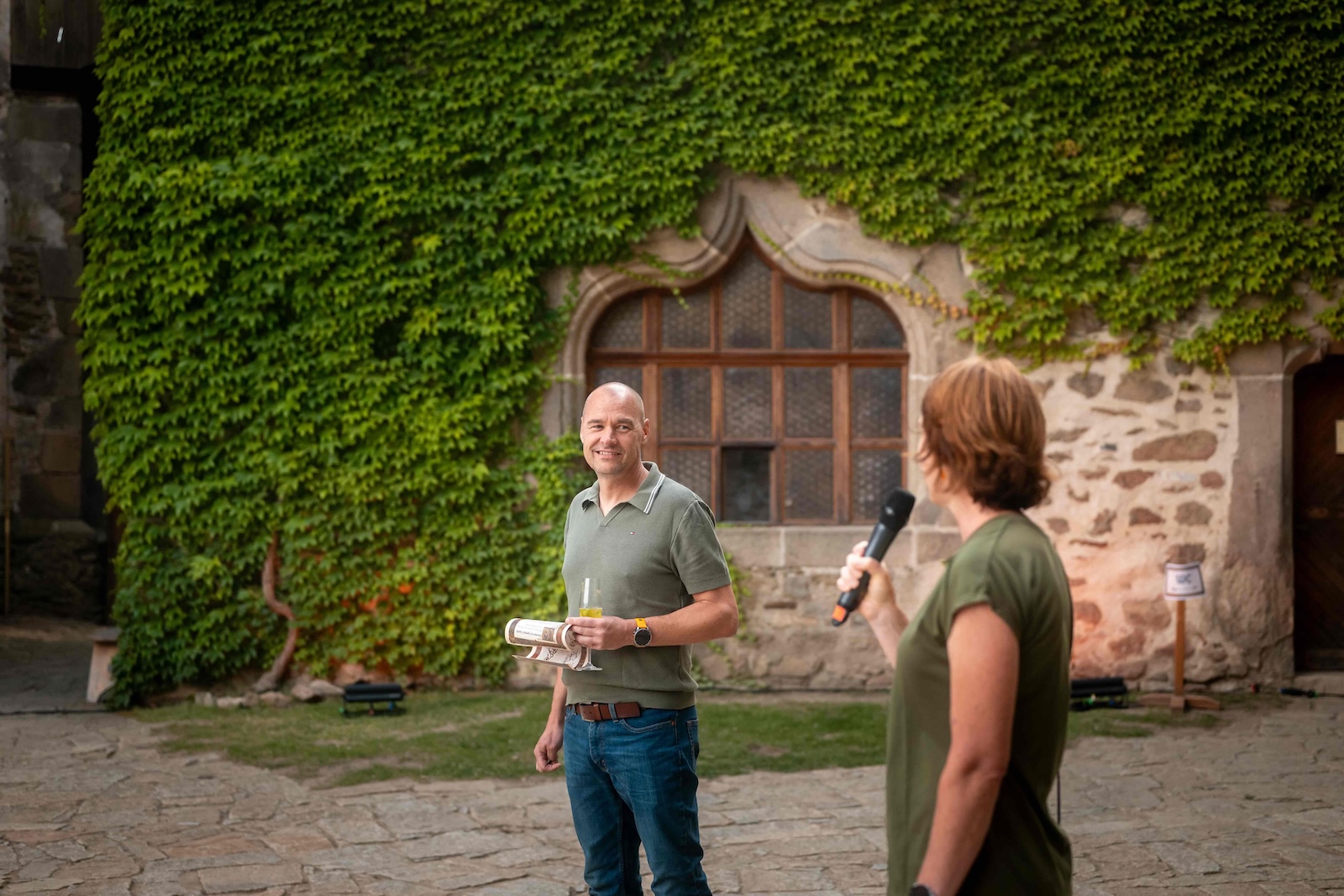 Host with a microphone talking to a guest in front of the ivy covered castle wall