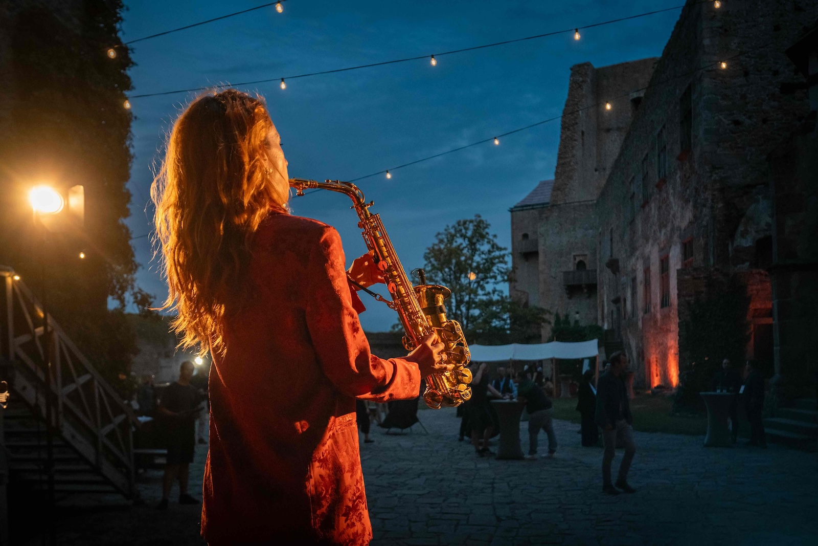 Saxophonist playing among the guests under string lights at dusk